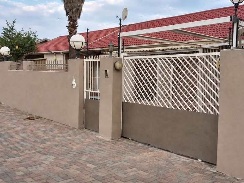 Front view of the property featuring a secure perimeter wall, sliding gate, pedestrian entrance, and paved driveway with exterior lighting.
