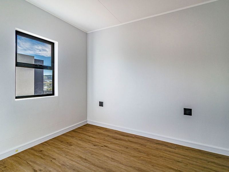 A bright, empty bedroom featuring modern laminate flooring, clean white walls, and a black-framed window providing natural light.