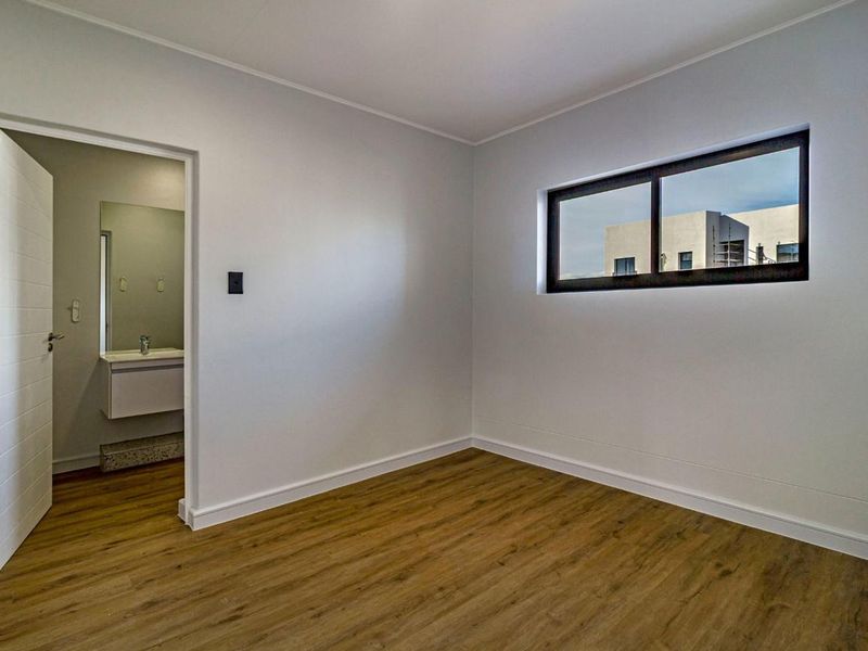 A bright, empty bedroom featuring modern laminate flooring, clean white walls, and direct access to an en-suite bathroom.