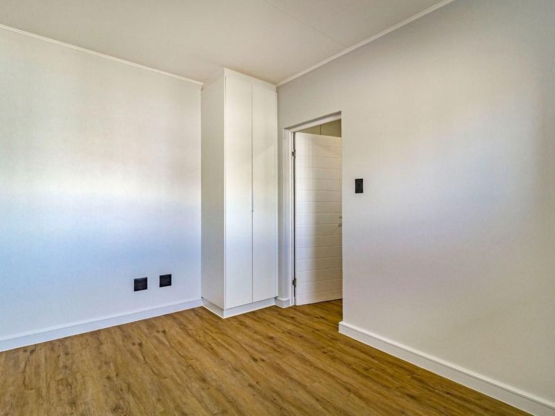 A bright, empty bedroom featuring modern laminate flooring, neutral walls, and a built-in cupboard, offering a clean and versatile living space.