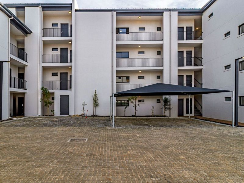 A modern apartment complex exterior featuring a paved parking area, shade cloth carport, and multi-level balconies with metal railings.