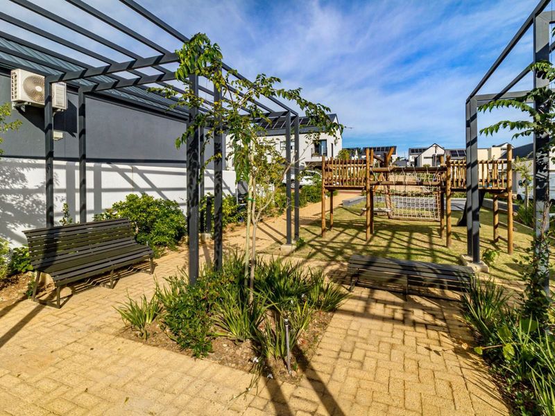 A well-maintained communal garden area featuring a pergola, paved pathways, and a wooden playground structure within a residential apartment complex.