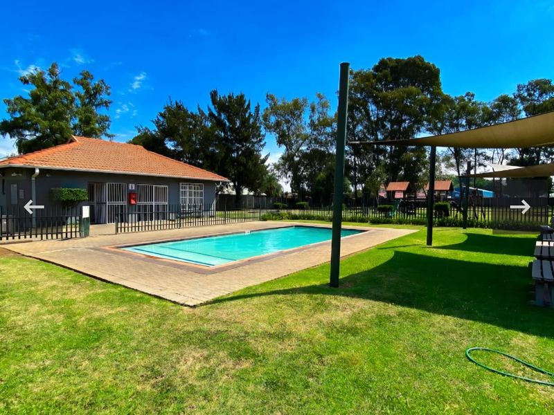 Communal swimming pool area featuring a paved deck, shade sails, and adjacent clubhouse building within a well-maintained garden setting.
