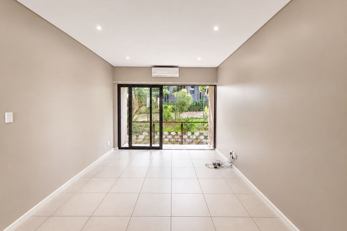 A bright, empty living room featuring neutral tiled flooring, recessed lighting, a split-system air conditioner, and sliding glass doors leading to an outdoor area.