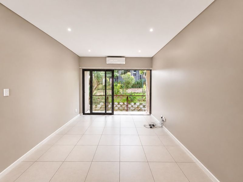 A bright, empty living room featuring neutral tiled flooring, recessed lighting, a split-system air conditioner, and sliding glass doors leading to an outdoor area.