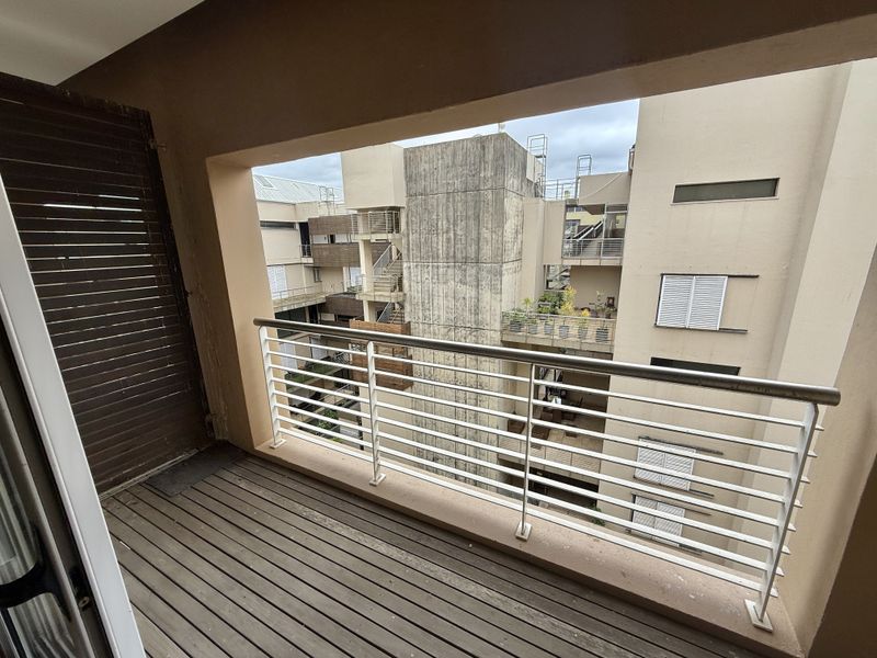 A covered balcony featuring timber decking and a metal balustrade, overlooking the apartment complex courtyard.