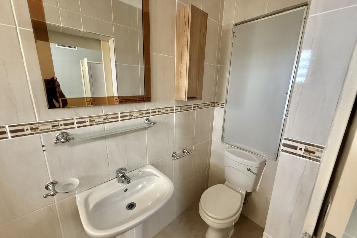 A clean, functional bathroom featuring a pedestal washbasin, toilet, wall-mounted storage cabinet, and neutral wall tiling with decorative accents.