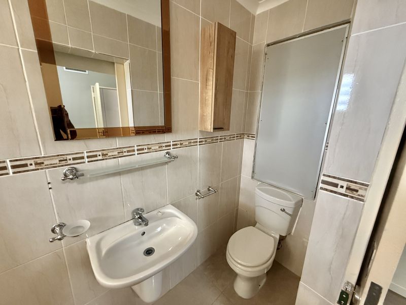 A clean, functional bathroom featuring a pedestal washbasin, toilet, wall-mounted storage cabinet, and neutral wall tiling with decorative accents.