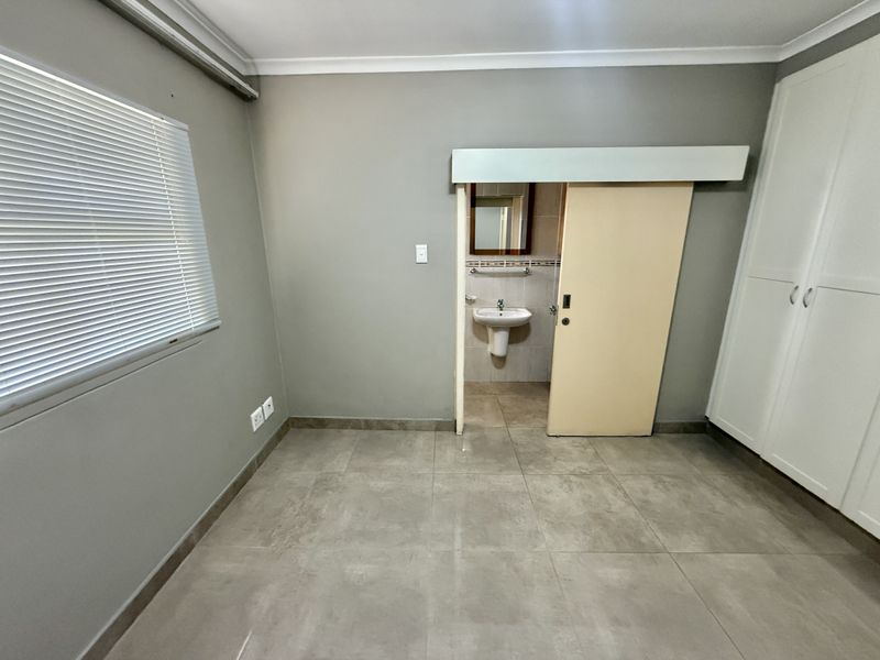 A spacious bedroom featuring tiled flooring, built-in cupboards, and an en-suite bathroom with a pedestal basin, presented in a clean, vacant condition.