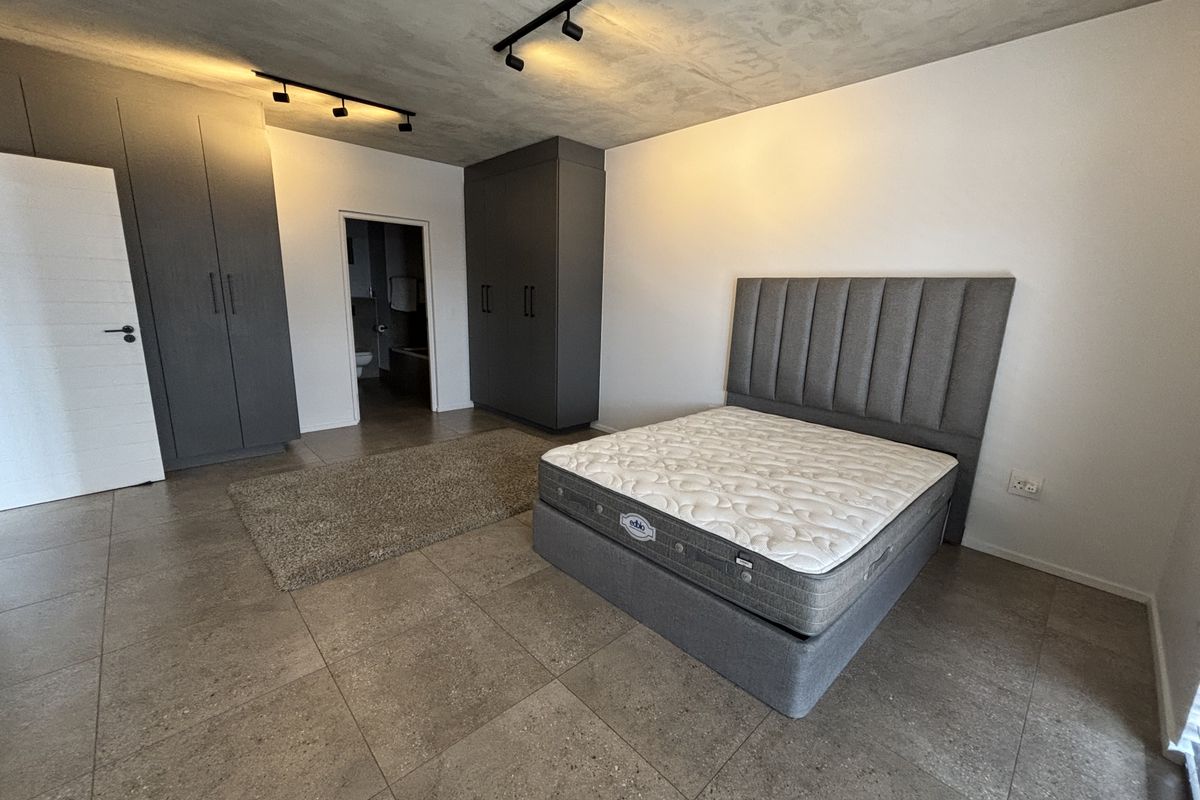 Spacious bedroom featuring modern exposed concrete ceilings, track lighting, large built-in cupboards, and tiled flooring with direct access to an en-suite bathroom.