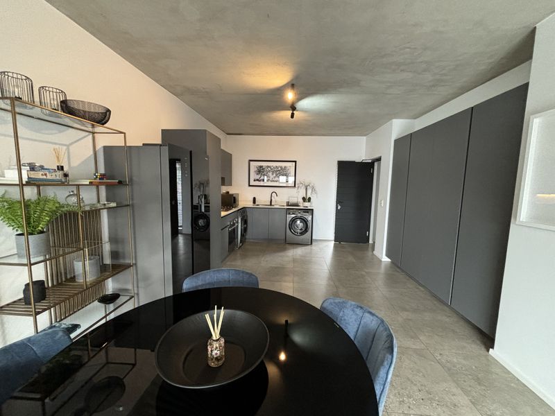 Modern open-plan kitchen and living area featuring sleek dark cabinetry, integrated appliances, tiled flooring, and a contemporary exposed concrete ceiling finish.
