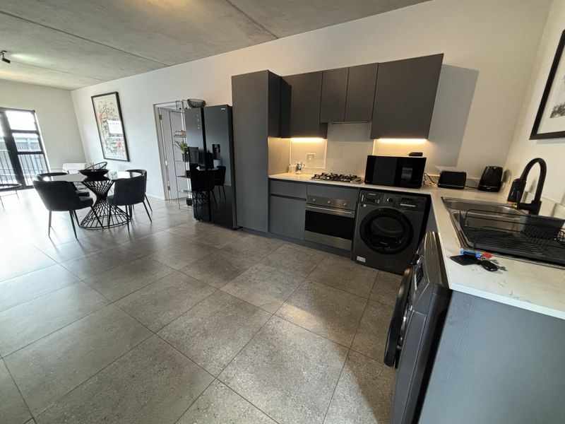 Modern open-plan kitchen featuring dark cabinetry, stone countertops, integrated appliances, and an industrial-style exposed concrete ceiling, flowing into the dining area.