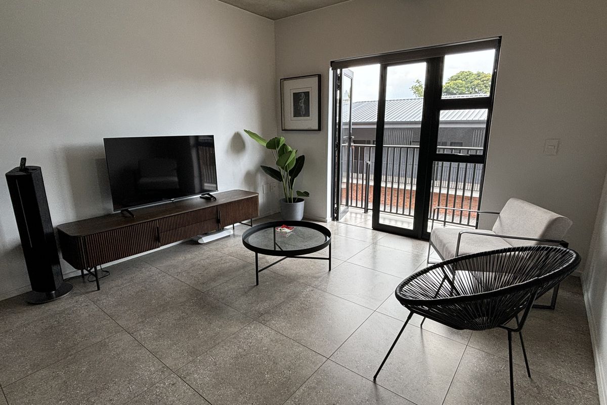 Modern living room featuring large format floor tiles, an exposed concrete ceiling, and direct access to a private balcony through black aluminium sliding doors.