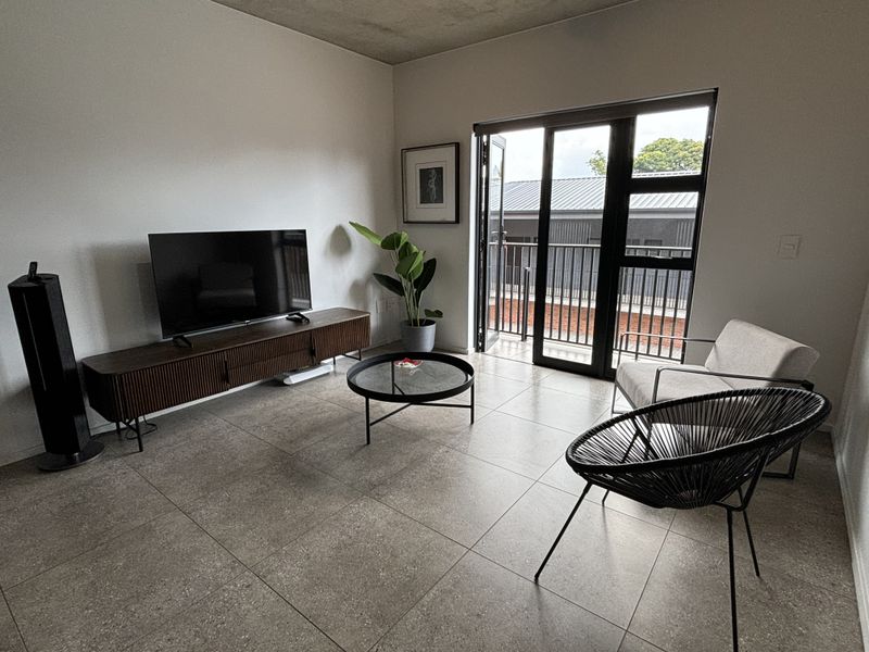 Modern living room featuring large format floor tiles, an exposed concrete ceiling, and direct access to a private balcony through black aluminium sliding doors.
