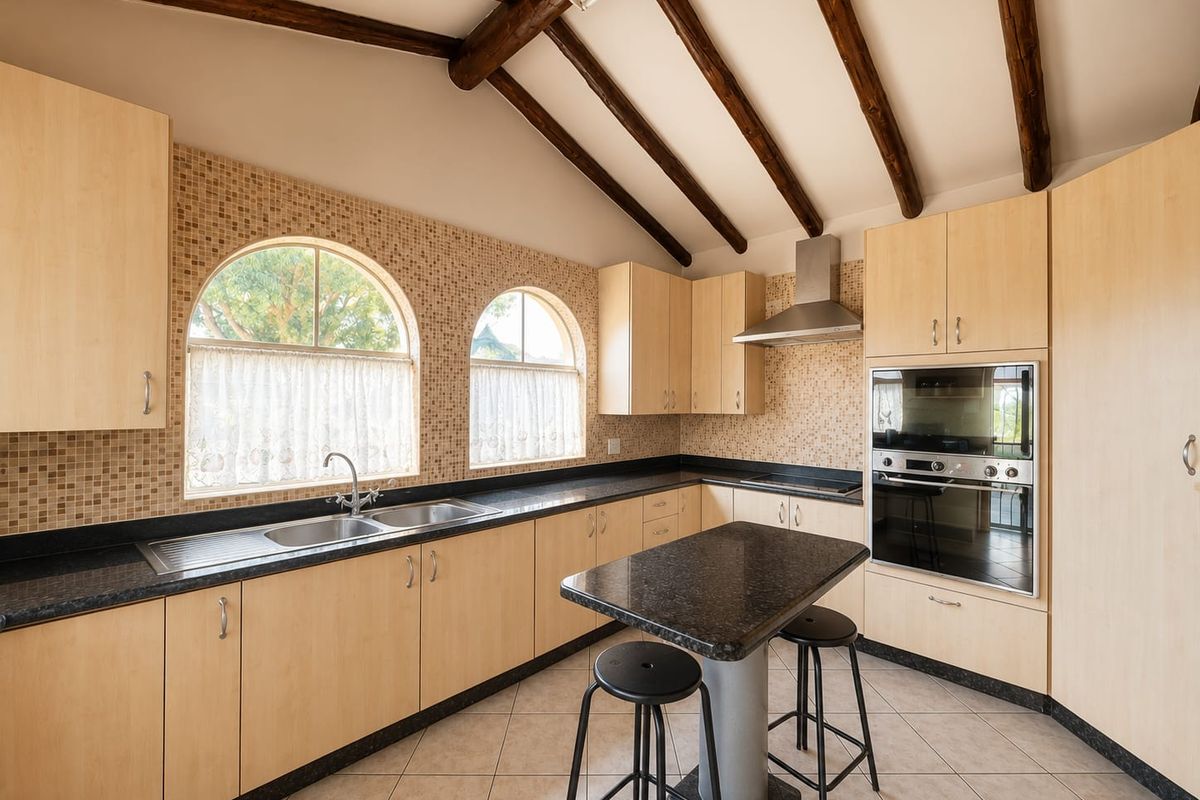 A spacious kitchen featuring exposed timber ceiling beams, arched windows, granite countertops, and built-in appliances with a central breakfast island.