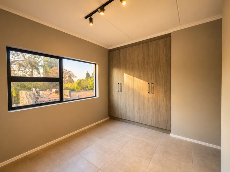 A bright, empty bedroom featuring modern tiled flooring, a large built-in wardrobe with wood-grain finish, and contemporary track lighting.