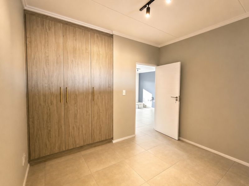 A clean, modern bedroom featuring neutral tiled flooring, built-in wood-finish cupboards, and contemporary track lighting, offering a bright and versatile living space.