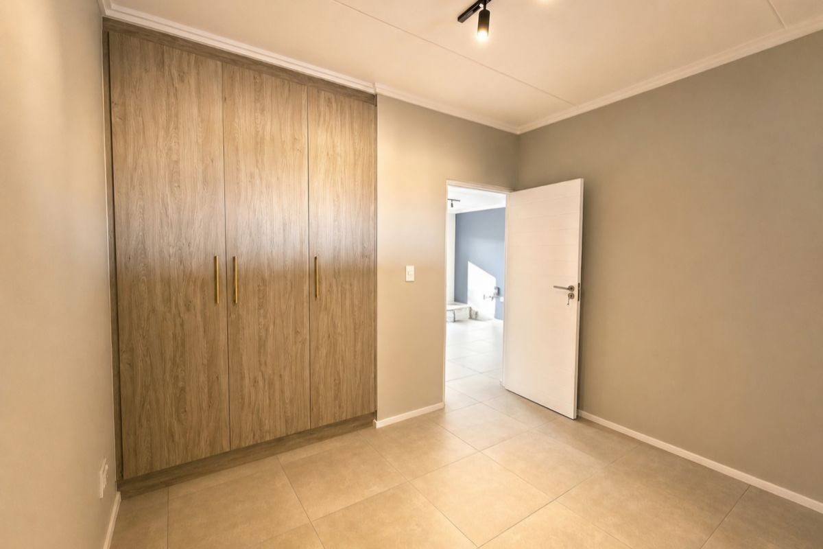 A clean, modern bedroom featuring neutral tiled flooring, built-in wood-finish cupboards, and contemporary track lighting, offering a bright and versatile living space.