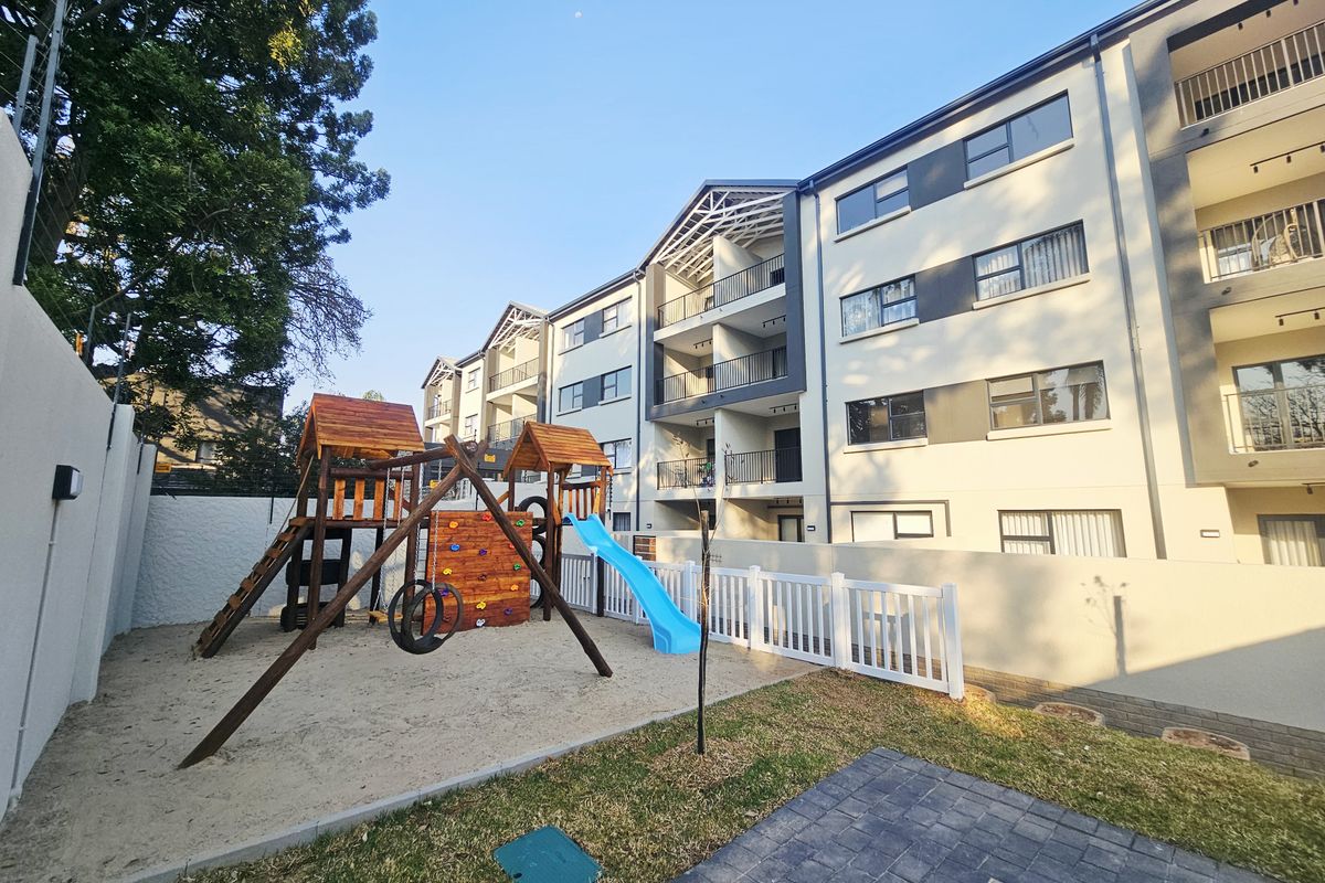Exterior view of an apartment complex featuring a communal playground area with sand pit, slide, and climbing frame, adjacent to paved walkways.