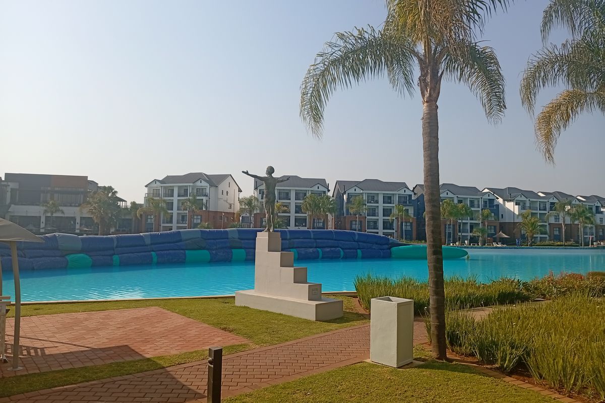 A well-maintained communal swimming pool area featuring landscaped gardens, paved walkways, and a central sculpture, set against a backdrop of modern apartment buildings.
