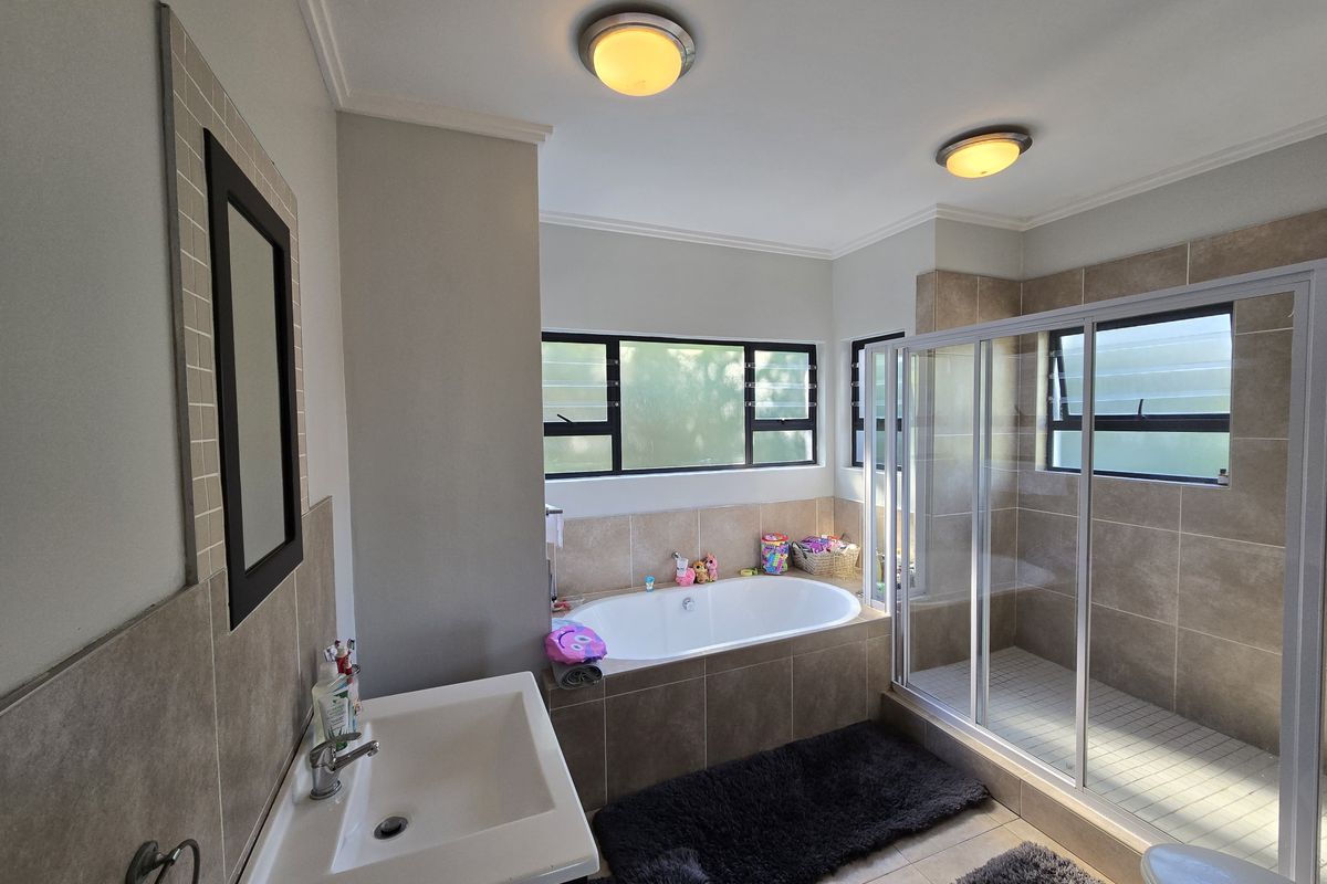 A well-maintained bathroom featuring a built-in bathtub, separate glass shower enclosure, vanity basin, and neutral wall tiling with natural light from louvre windows.
