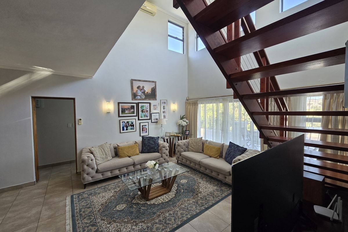 Spacious double-volume living room featuring tiled floors, a prominent wooden staircase, and large windows that provide ample natural light.