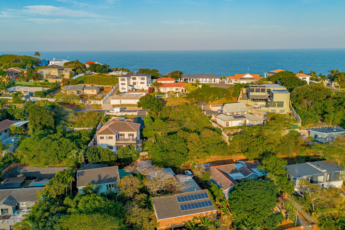 An aerial view of a coastal residential neighborhood featuring various homes with tiled roofs and solar panel installations set within a lush green environment.