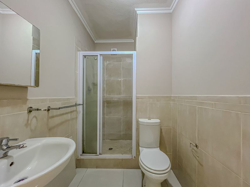 A functional bathroom featuring a shower enclosure, pedestal basin, and toilet with neutral wall and floor tiling.