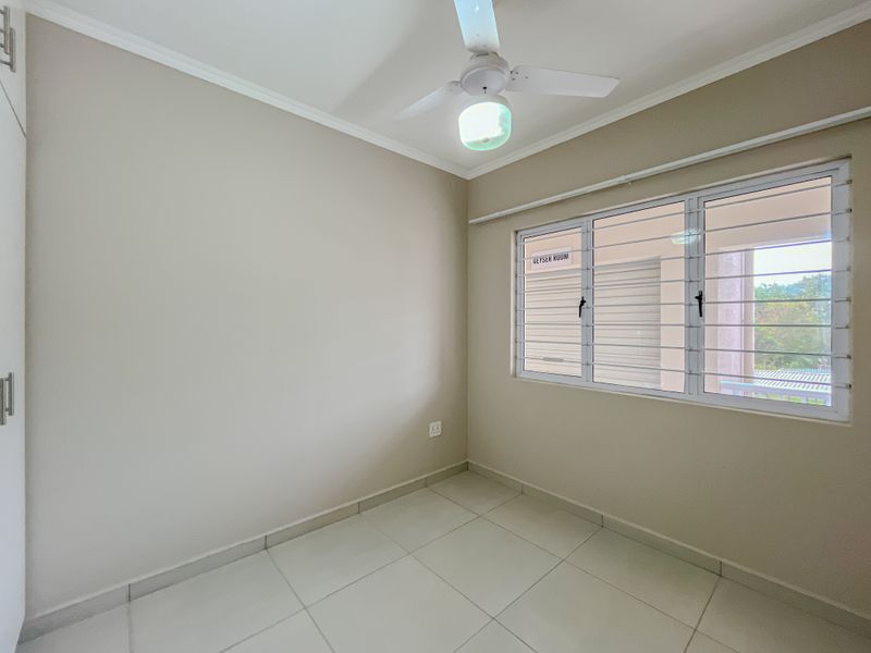 A clean, empty bedroom featuring neutral tiled flooring, built-in cupboards, a ceiling fan, and a window fitted with security bars.