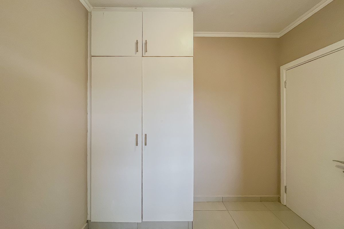 A clean, empty bedroom featuring neutral wall tones, tiled flooring, and a white built-in wardrobe with overhead storage.