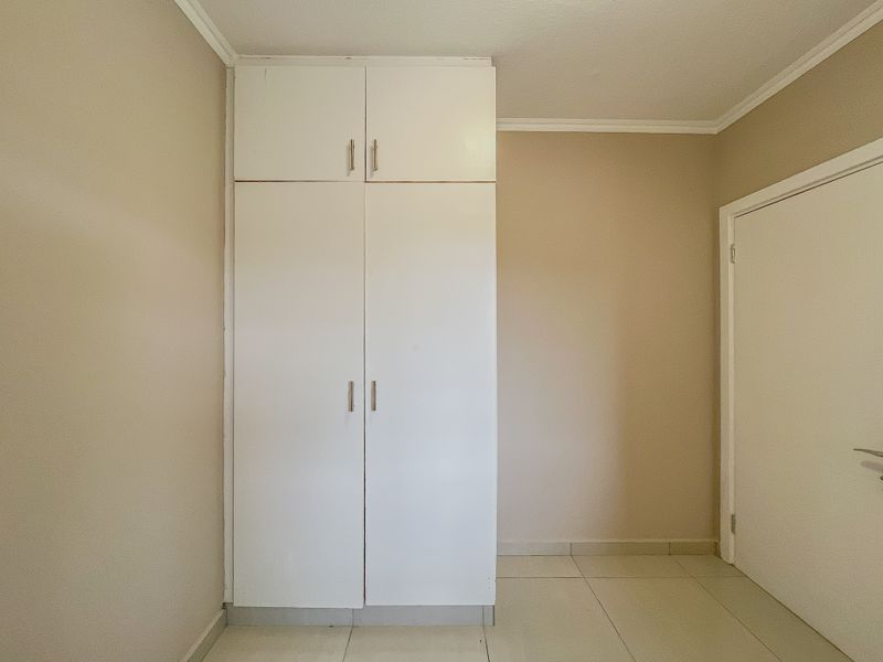 A clean, empty bedroom featuring neutral wall tones, tiled flooring, and a white built-in wardrobe with overhead storage.