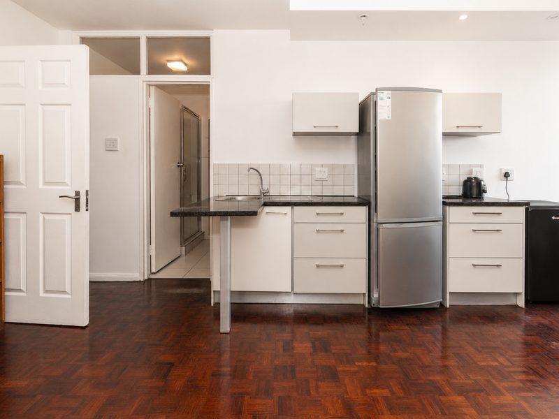 Compact kitchen featuring white cabinetry, granite countertops, tiled backsplash, and warm parquet flooring, with an adjacent doorway leading to a bathroom.