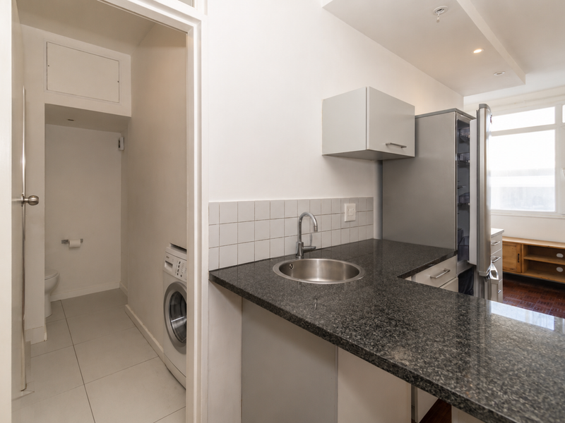 Compact kitchen area featuring granite countertops, stainless steel sink, and tiled backsplash, with an adjacent utility space containing laundry plumbing.