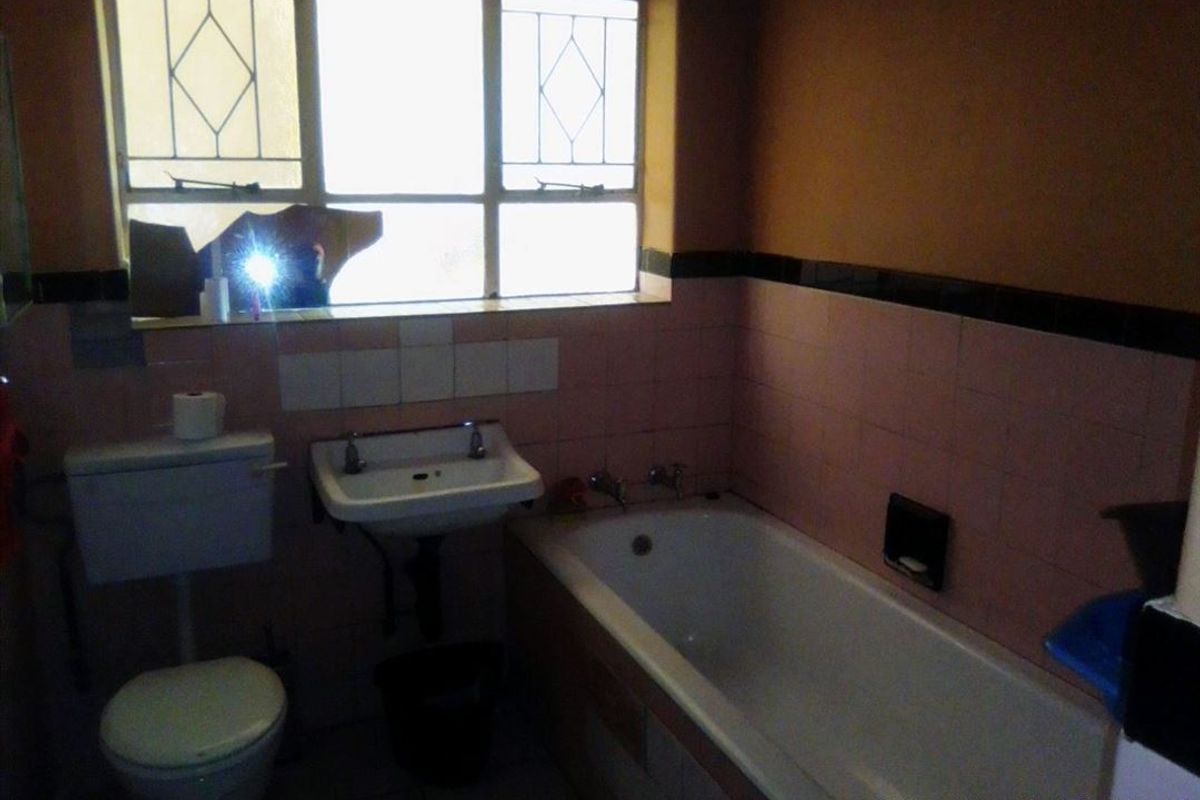 A dated bathroom featuring a bathtub, wall-mounted basin, and toilet, with pink wall tiles and a broken window pane requiring repair.