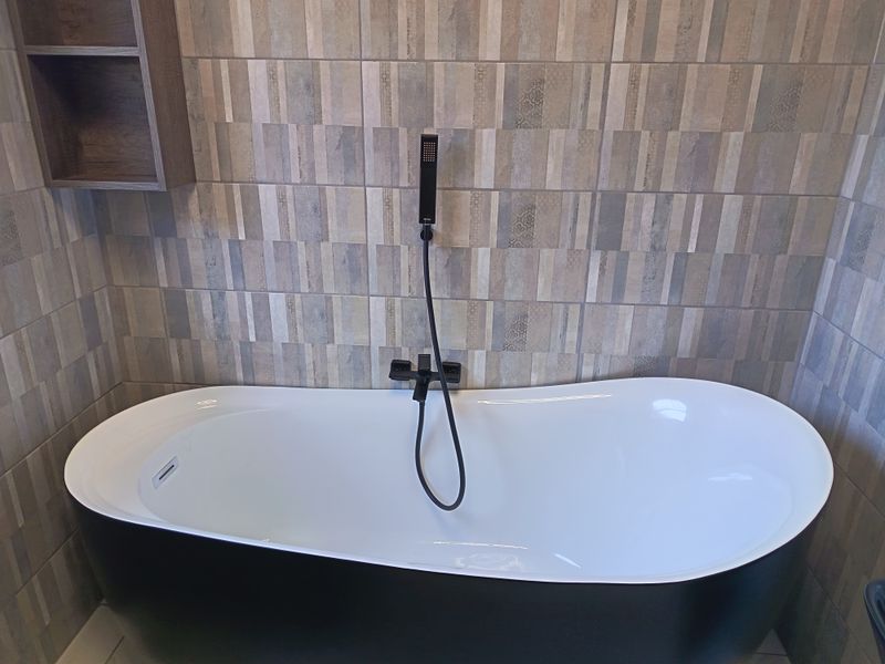 Modern bathroom featuring a sleek black and white freestanding bathtub with a contemporary matte black hand shower and decorative tiled wall finish.