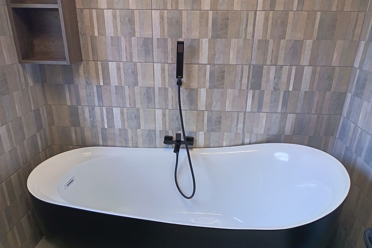 Modern bathroom featuring a sleek black and white freestanding bathtub with a contemporary matte black hand shower and decorative tiled wall finish.