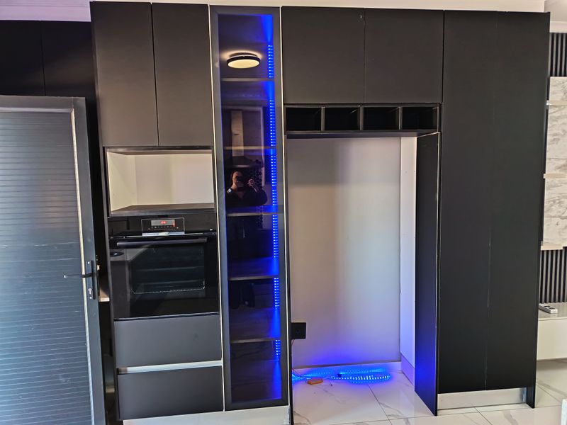Modern kitchen wall featuring matte black cabinetry, a built-in oven, integrated shelving with LED accent lighting, and a dedicated refrigerator alcove.