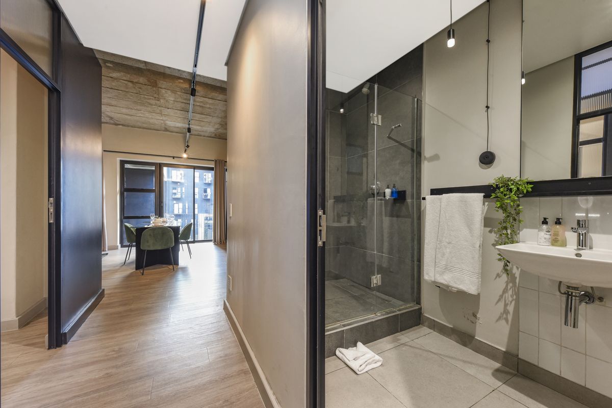 Modern bathroom featuring a walk-in shower with glass screen, wall-mounted basin, and contemporary finishes, adjacent to an open-plan living area with exposed concrete ceilings.