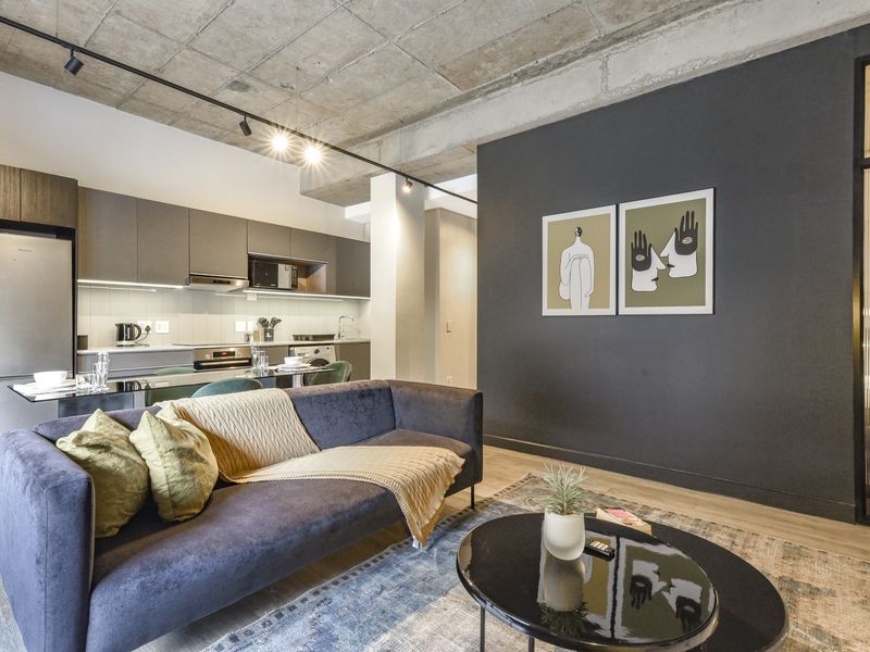 Modern open-plan living area featuring an exposed concrete ceiling, track lighting, and a sleek, well-appointed kitchen with integrated appliances and contemporary finishes.