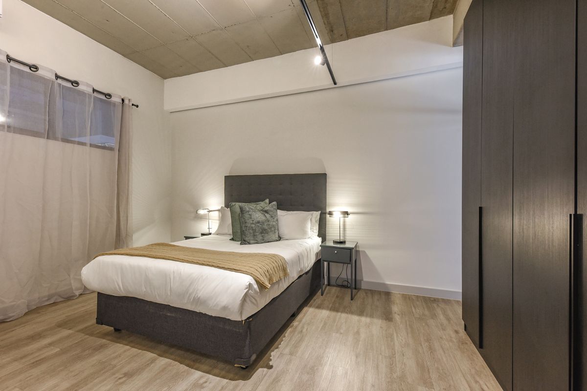 Modern bedroom featuring an exposed concrete ceiling, track lighting, laminate flooring, and a large built-in wardrobe.