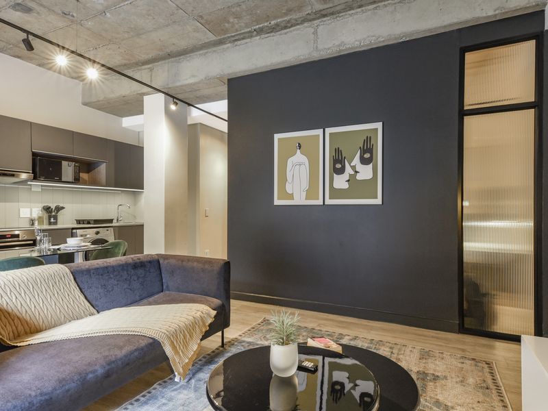 Modern open-plan living area featuring an exposed concrete ceiling, track lighting, and a sleek kitchen with integrated appliances and contemporary finishes.
