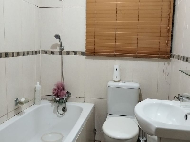 A compact bathroom featuring a bathtub with shower mixer, pedestal basin, and toilet, finished with neutral wall tiles and a window blind.