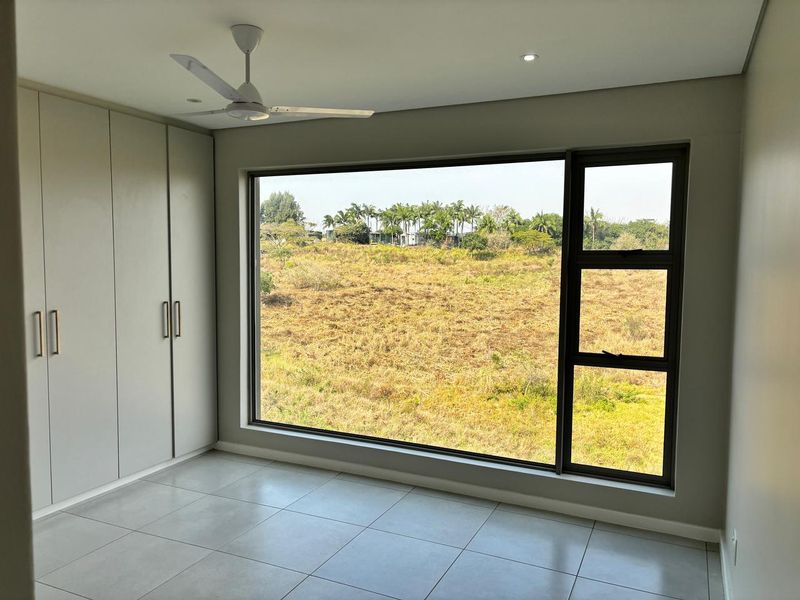 A bright, modern bedroom featuring neutral tiled flooring, floor-to-ceiling built-in cupboards, a ceiling fan, and a large window offering expansive natural views.