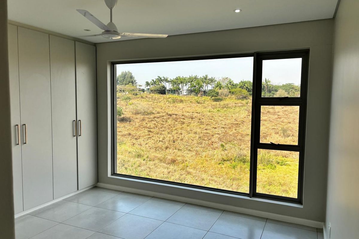 A bright, modern bedroom featuring neutral tiled flooring, floor-to-ceiling built-in cupboards, a ceiling fan, and a large window offering expansive natural views.