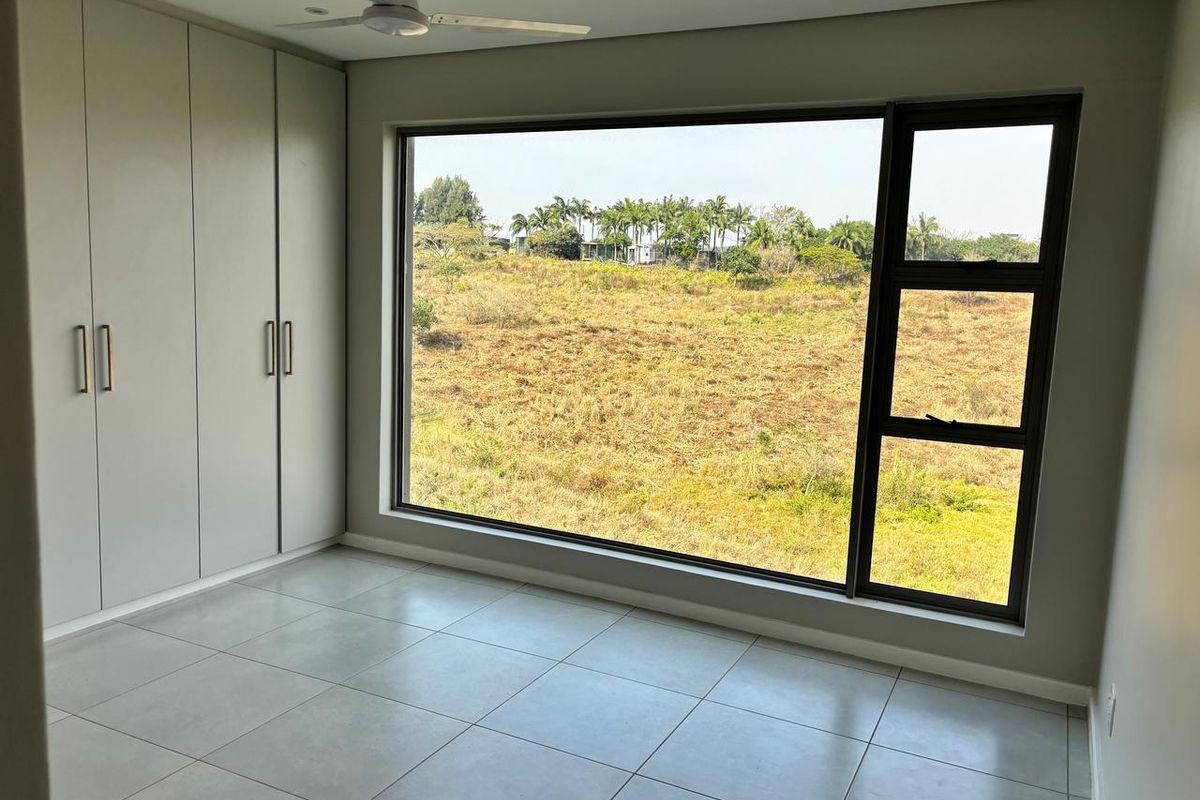 A bright, modern bedroom featuring large aluminium windows with scenic views, neutral tiled flooring, and ample built-in cupboard space.