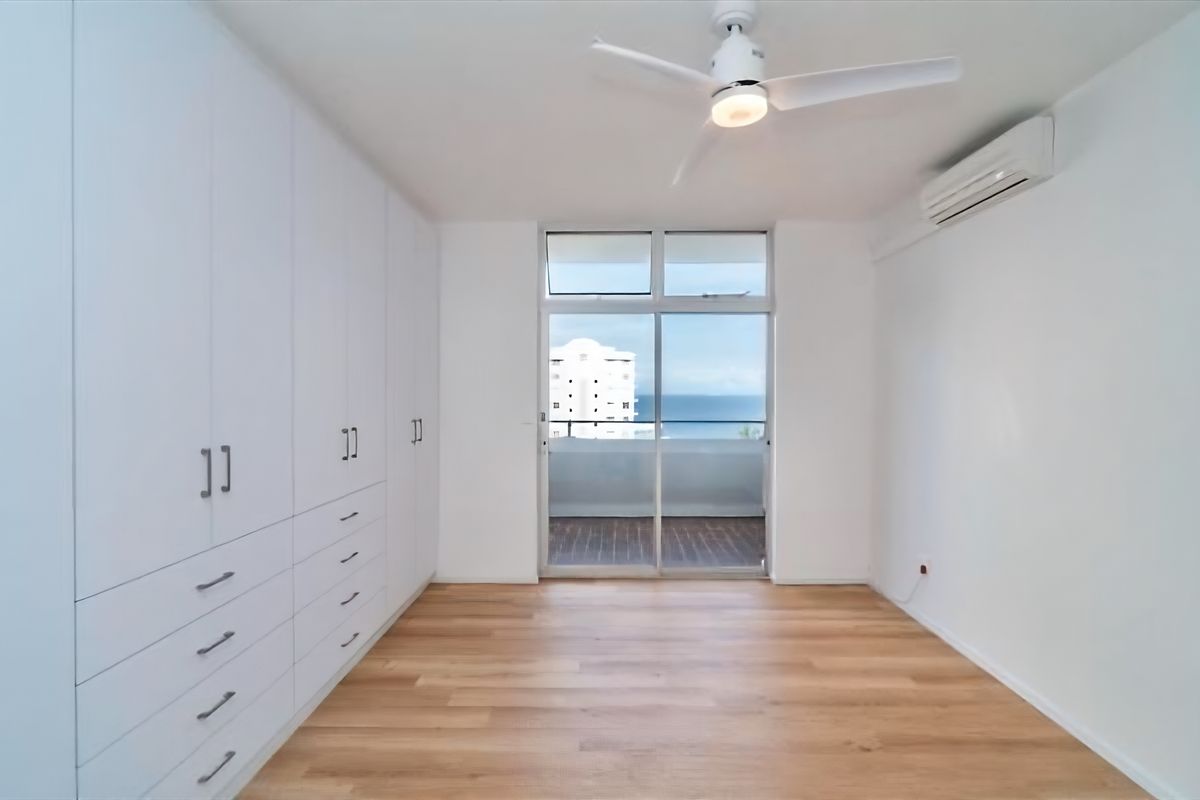 A bright, empty bedroom featuring extensive built-in cabinetry, laminate flooring, a ceiling fan, air conditioning, and direct access to a balcony with ocean views.