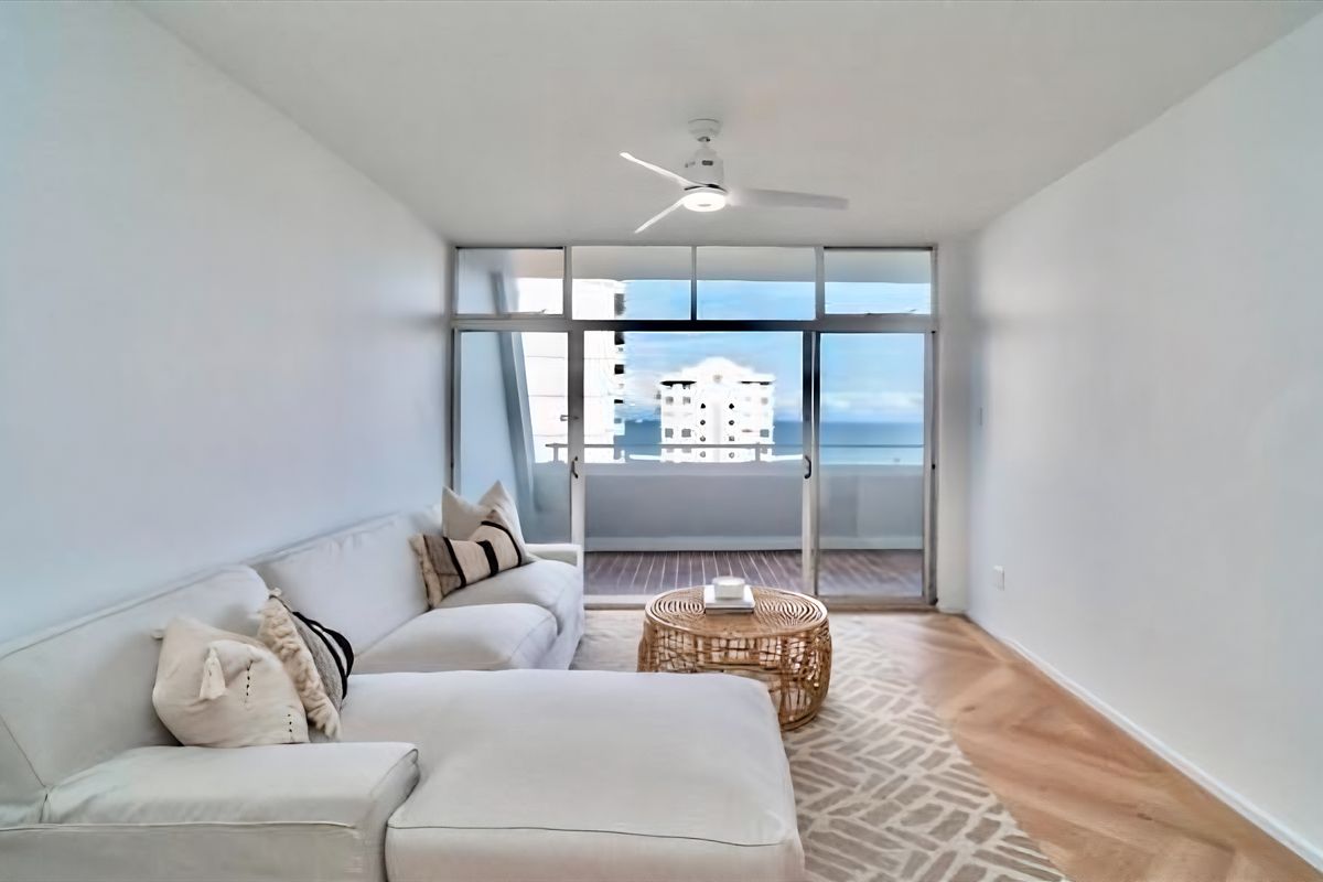 Bright living room featuring herringbone timber flooring, a modern ceiling fan, and large sliding glass doors opening to a balcony with coastal views.