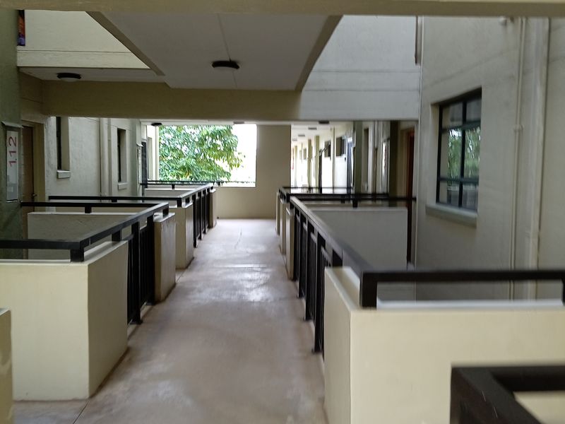 An open-air apartment complex hallway featuring concrete flooring, metal safety railings, and recessed lighting, providing access to residential units.