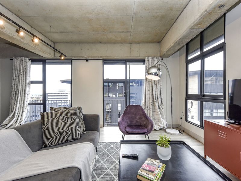 Modern living room featuring an industrial-style exposed concrete ceiling, track lighting, and large windows providing ample natural light and urban views.