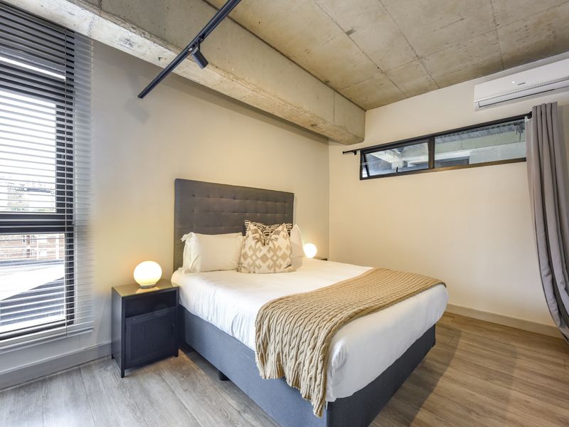 Modern bedroom featuring an exposed concrete ceiling, laminate flooring, split-system air conditioning, and large windows with venetian blinds.